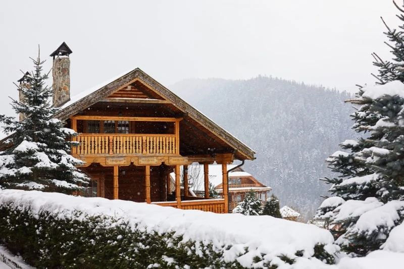 Christmas wooden mansion in mountains on snowfall winter day. Cozy chalet on ski resort near pine forest. Cottage of round timber with wooden balcony. Fir-trees covered with snow. Chimneys of stone.